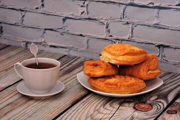 A cup of black tea with a spoon on a saucer and a plate with an assortment of homemade cakes on a wooden table against a brick wall.