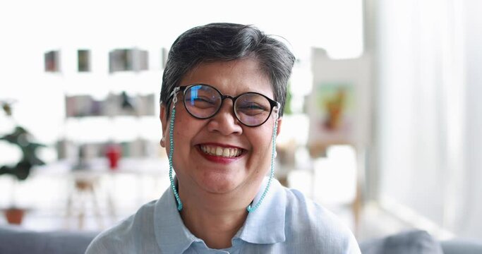 Middle aged Asian female in glasses looking at camera and laughing in light room at home