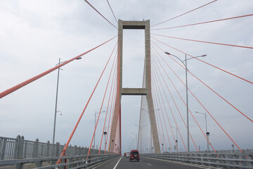Scene of the famous Suramadu Bridge and its red suspension steel cables with cars on road and cloudy sky background. 