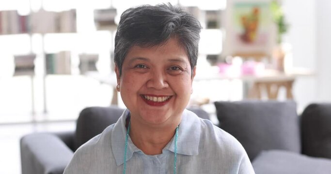 Optimistic mature Asian woman with short hair looking at camera and laughing at joke in sunlit room