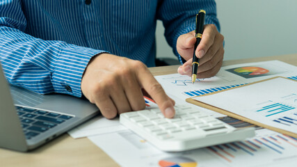 A businessman or accountant holds a pen and presses a calculator to calculate business information. Financial accounting graph document and notebook computer in the company office.