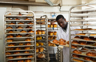 Portrait of african american baker working in bakehouse, putting tray with fresh bakery goods on...