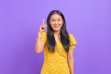 Smiling young Asian woman pointing fingers up and having an idea on purple background