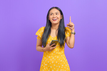 Smiling young Asian woman using a mobile phone and pointing fingers up with an idea on purple background