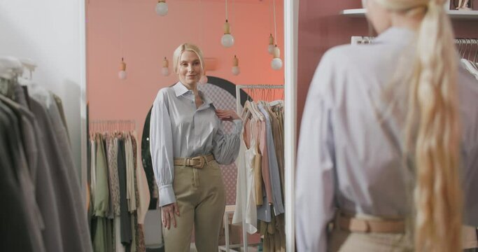 Beautiful Young Blonde Woman Trying on Jacket in Front of Mirror. Lady Shopping Choosing Dresses Looking in Mirror Uncertain. Beautiful Young Multicultural Shopper in Clothing Store.
