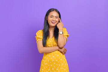 Smiling young Asian woman wearing yellow dress and looking confident on purple background