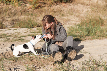 Naklejka na ściany i meble young beautiful brown haired woman sitting on a sandy beach and playing with a dog. jack russel terrier grabbing a toy and trying to take it away. autumn leisure with a dog, playful pets