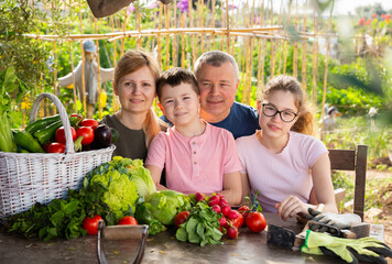 Cheerful family of four sitting at wooden table in garden on summer day happy with rich harvest of vegetables.