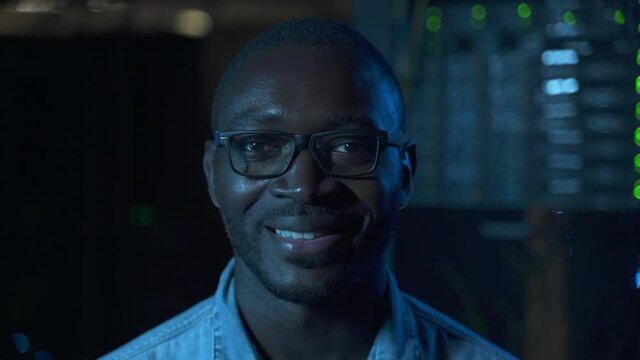 IT engineer in data center. Skilled African-American system administrator with glasses smiles to camera spbas standing against operating servers closeup