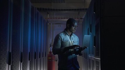 Engineer IT technician sets up system. Young African-American specialist in shirt spbas fixes operating equipment with contemporary laptop in corridor closeup