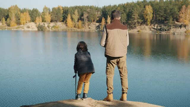 Back view of adult man and little boy fishing in lake on sunny autumn day enjoying time together and beautiful nature. Family and leisure activities concept.
