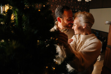 Loving young couple decorating Christmas tree with shiny balls standing hugging near fireplace in cozy dark living room. Happy man and woman preparing home for New Year holidays celebration