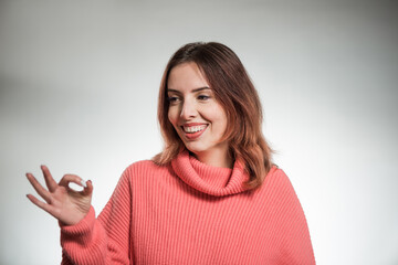 young positive woman in pink sweater showing ok sign on studio background