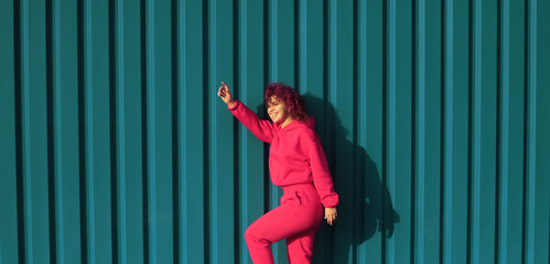 Positive woman with curly pink hair in magenta hoodie posing outdoors. Smiling happy woman concept. Beautiful curly hair woman on blue background. 