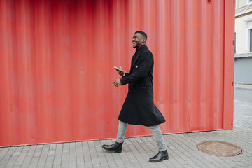 Overjoyed black man in a warm coat walking in front of corrugated wall