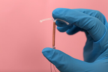 Doctor holding T-shaped intrauterine birth control device on pink background, closeup. Space for...