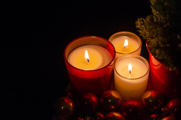 Selective focus on flame burning of red glass candle with Christmas tree and bauble ornaments in dark background.