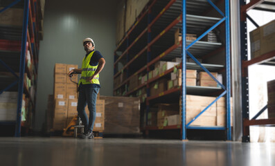 Handsome young male employee concentrating and focusing on work wearing safety helmet and uniform in warehouse while standing between racks in aisle