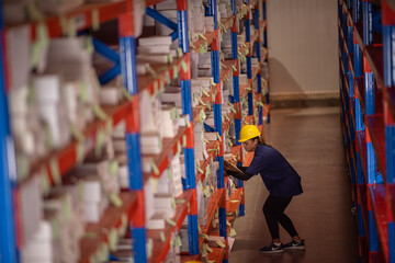 Young African-American woman wearing uniform and hardhat helmet working while checking inventory in aisle using clipboard for cross-verification and updating records