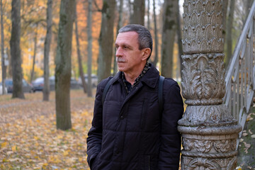 Fototapeta na wymiar Senior retired man on beautiful autumn day while walking in park stands and rests by old column and deeply thought.