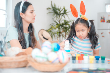 Obraz na płótnie Canvas Beautiful young Asian woman wearing rabbit hairband painting eggs using paint colors in a creative work to celebrate easter with cute little daughter at home