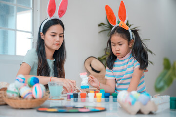 Beautiful young Asian woman wearing rabbit hairband painting eggs using paint colors in a creative work to celebrate easter with cute little daughter at home