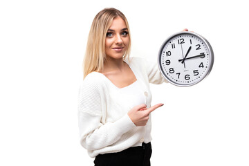 Photo of a beautiful pretty positive smiling young blonde woman with makeup in a stylish white knitted sweater and black pants points her finger at the big clock isolated on a white background with