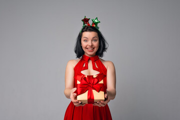 Happy young brunette woman in red dress posing on gray background and showing gift box decorated with red ribbon. Holidays, celebration and lifestyle concept