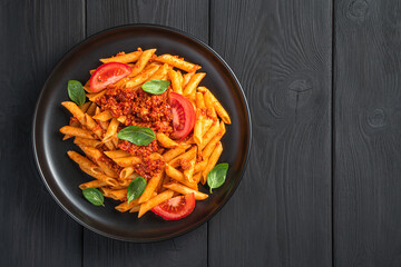 Penne pasta with bolognese sauce and basil in a black plate on a black background.