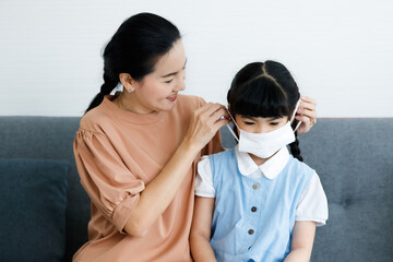 Close up shot of Asian lovely mother sitting on sofa in living room helping little cute daughter in school uniform wearing safety hygiene protecting face mask preventing coronavirus during outbreak