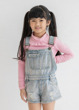 Portrat closeup studio shot of Asian happy little cute preschooler girl with pigtail hair wearing pink long sleeve shirt with jeans overalls standing posing smiling look at camera on white background