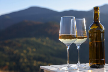 Glasses and bottle of white wine on wooden table against mountain landscape, space for text