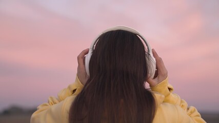 put on headphones on your head, a beautiful pink sunset in sky, listen to music tracks in speakers with your ears, get musical pleasure and relax, record audio at sunset, diversify your leisure time
