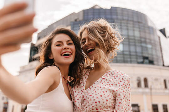 Close-up image of two female friends taking photo on modern smartphone on background of building. Brunette and blonde are smiling broadly with teeth, dressed in light loose clothes.