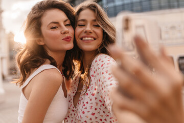 Sweet young european women pose for selfie against blurred city background. Two funny beauties with wavy hair are dressed in summer casual clothes on sunny day.