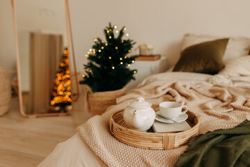 Christmas concept. A teapot of tea and a mug on a wicker tray stand on the bed in a cozy decorated bedroom at home on a New Year's holiday