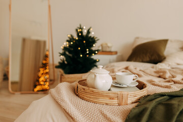 Christmas concept. A teapot of tea and a mug on a wicker tray stand on the bed in a cozy decorated bedroom at home on a New Year's holiday