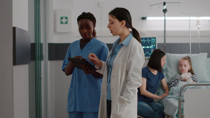 Obraz premium African american physician nurse checking disease expertise on tablet discussing therapy recovery with pediatrician doctor working in hospital ward. Sick child resting in bed after medical surgery