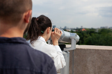 Fototapeta na wymiar Husband surprising wife with romantic panoramic view of metropolitan city celebrating relationship anniversary. Woman looking through binocular telescope from observation point