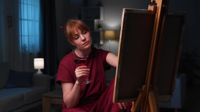 Beautiful woman artist draws a picture of the house and drinks red wine from the glass.