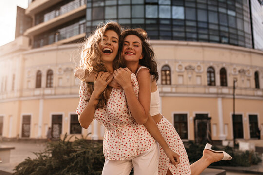 Two funny young women go crazy outside during warm season. Blonde with brunette with snow-white smile embracing each other, dressed in casual light clothes. Vacation, happiness and pleasure concept