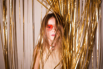 stylish model baby child girl in pink glasses on background of golden tinsel. concept of holiday,...