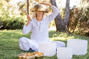 Woman in white clothes sit on green grass in park. Person hold metal singing bowl on head and make...