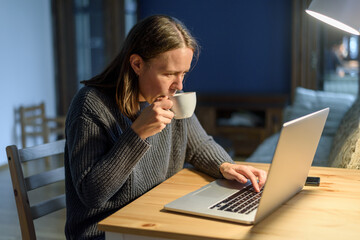 Woman in knitted sweater working on laptop at home living room. Drinking coffee or tea and typing