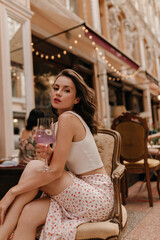 Young modern woman sits on chair sipping cocktail at street cafe. Cute caucasian beauty dressed in...