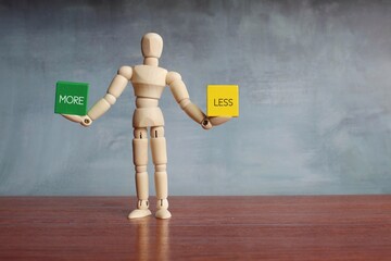 Wooden human figure balancing wooden cubes with text MORE and LESS.