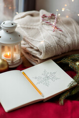 christmas, holidays and objects concept - close up of sketchbook with pencil drawing of snowflake, warm wool braided sweater, lantern and fir branch on red tablecloth on window sill at home