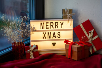 holidays, decoration and celebration concept - close up of merry christmas greeting on light box and gift boxes on red tablecloth on window sill at home