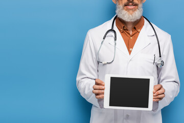 cropped view of bearded middle aged doctor in white coat holding digital tablet with blank screen on blue