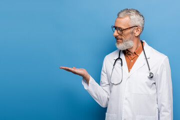 bearded middle aged doctor in glasses and white coat pointing with hand on blue